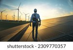 Professional Male Green Energy Engineer Walking On Industrial Solar Panel, Wearing Safety Belt And Hard Hat. Man Inspecting Sustainable Energy Farm With Wind Turbines On Background.