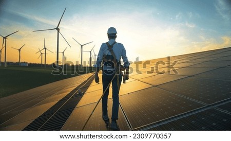 Professional Male Green Energy Engineer Walking On Industrial Solar Panel, Wearing Safety Belt And Hard Hat. Man Inspecting Sustainable Energy Farm With Wind Turbines On Background.
