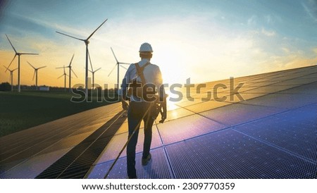 Professional Male Green Energy Engineer Walking On Solar Panel, Wearing Safety Belt And Hard Hat. Man Inspecting Sustainable Energy Farm With Wind Turbines. VFX Edit Visualizing Electricity Flow.