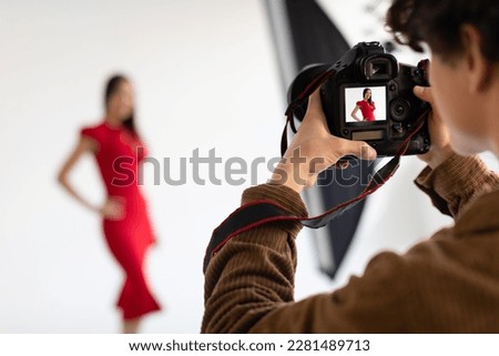 Professional male photographer and beautiful female model in red dress making beauty or content photoshoot in photostudio on white background, focus on camera