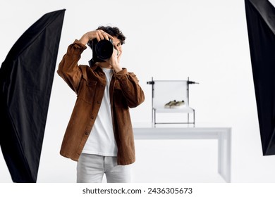 Professional male photographer in corduroy jacket focusing his DSLR camera, preparing to take photograph in well-lit studio with soft boxes, free space - Powered by Shutterstock