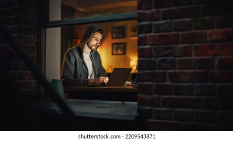 Professional Male Programmer Writing Code For Complex Project And Working Remotely On Laptop From Home Office. Freelance Software Engineer Designs App From Apartment. View From Outdoors Into Window.