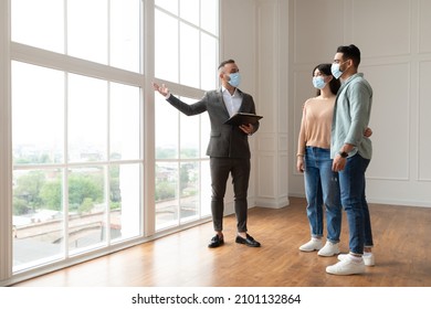 Professional Male Real Estate Agent In Medical Mask Showing Property House For Sale Or Rent To Young Married Couple, Holding Clipboard. New Home, Moving And Mortgage Loan. People Looking Out Of Window