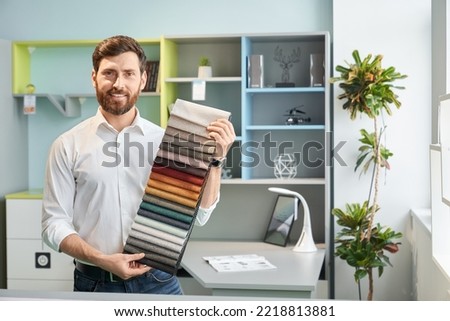 Professional male sales manager demonstrating various samples of upholstery fabric in light showroom. Portrait of bearded designer showing colorful palette, while smiling. Concept of interior design. Foto d'archivio © 