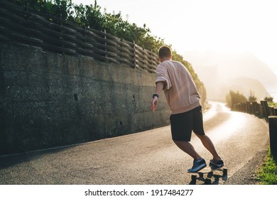 Professional male skater in casual wear training on board during evening sunset with orange light, handsome professional sportsman enjoying freestyle riding on longboard during vacations in summer