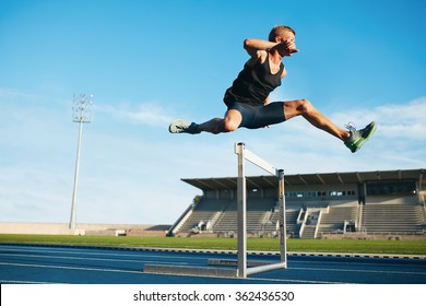 Professional male track and field athlete during obstacle race. Young athlete jumping over a hurdle during training on racetrack in athletics stadium. - Powered by Shutterstock