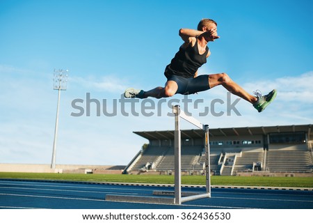 Professional male track and field athlete during obstacle race. Young athlete jumping over a hurdle during training on racetrack in athletics stadium.
