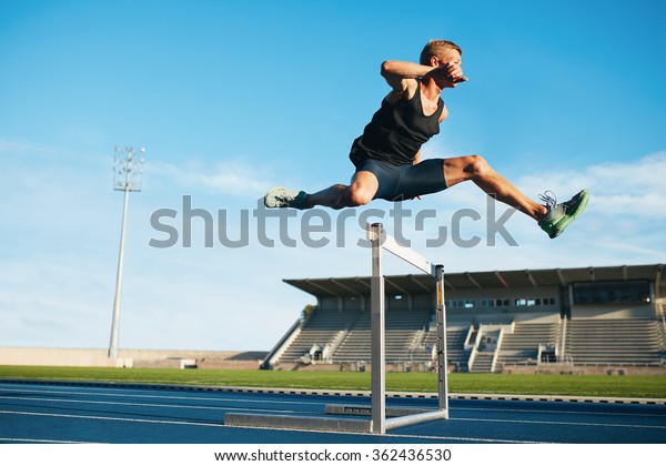 Professional male track and field athlete during\
obstacle race. Young athlete jumping over a hurdle during training\
on racetrack in athletics\
stadium.