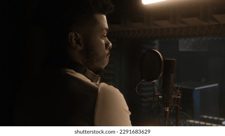 Professional male vocalist prepares to record cool track in front of microphone in soundproof room. African American singer works in sound recording studio. Concept of music production. Back view.