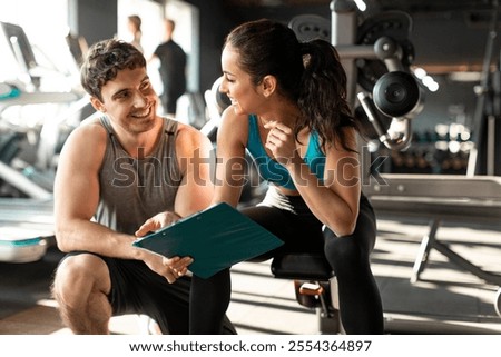 Professional man personal trainer explaining exercises with clipboard to female client, sitting in gym and talking