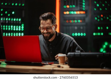 Professional man smiling while working on a laptop in a data center, managing server racks. Focusing on cybersecurity