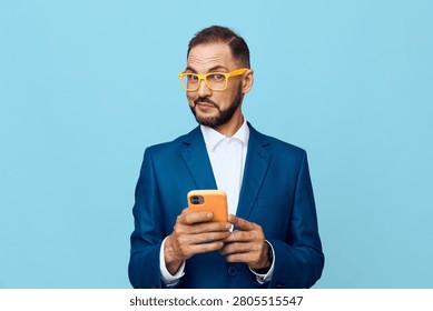 A professional man stands against a solid blue background, wearing a blue suit and yellow glasses, focused on his phone, expressing confidence and curiosity with a calm, composed demeanor throughout