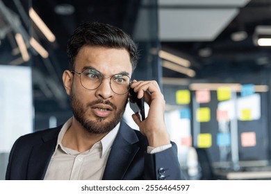 A professional man in a suit communicates on a smartphone, expressing focus and concern in a modern office setting. His intense expression indicates engagement in an important conversation. - Powered by Shutterstock