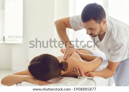 Professional masseur or manual therapist massaging young woman's shoulder, easing pain and relaxing muscles. Female patient getting remedial body massage in health clinic or physiotherapy center