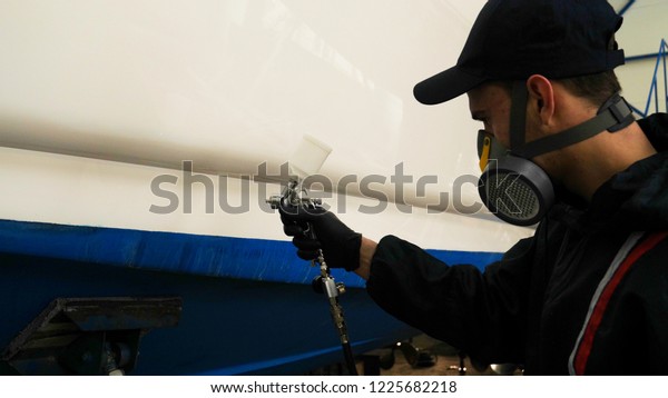 Professional master man in a robe and puller\
with spray gun puts a layer of paint or ceramics on the yacht.\
Concept of: Dangerous work, Chemistry, Paints, Garage, Transport\
Service.