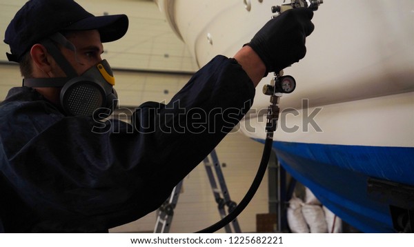 Professional master man in a robe and puller\
with spray gun puts a layer of paint or ceramics on the yacht.\
Concept of: Dangerous work, Chemistry, Paints, Garage, Transport\
Service.