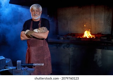 Professional mature blacksmith protective apron and gloves posing at workplace. Caucasian man with grey beard and hair standing with crossed arms at forge.  - Powered by Shutterstock