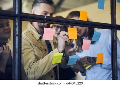 Professional matured coach writing on stickers ideas of strategy improvement supervising work of staff members,skilled team leader making notes of task while organizing working process in office 