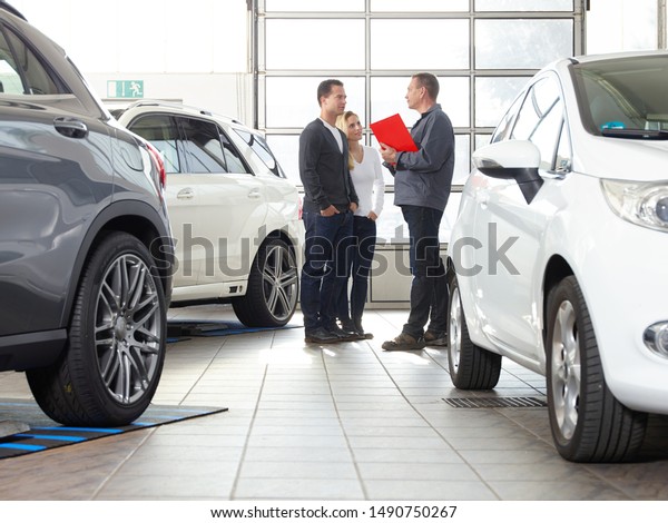 a professional mechanical car and tire service\
employee in his workwear stand with a pair of a blonde women and\
his husband in the garage between cars and give them consulting to\
the workshop service
