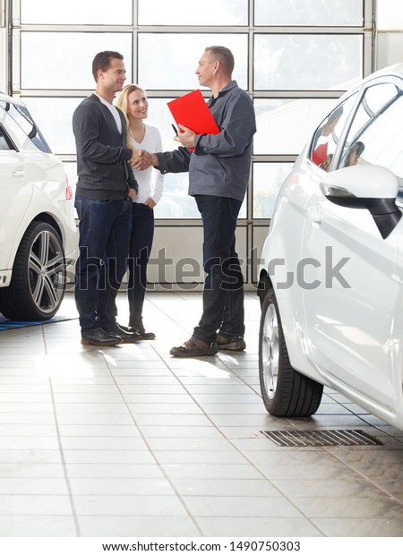 a professional mechanical car and tire service\
employee in his workwear stand with a attractive pair of a blonde\
women and his husband in the garage between cars and give handshake\
for a good deal