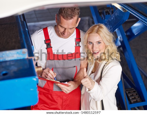 the professional mechanical employee of the car\
service workshop stand with the blonde female customer in the\
garage under her car and make a check list, she show thumb up for\
agree and smile