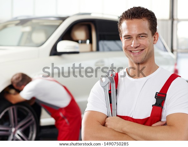 a professional mechanical employee of a car\
service workshop stand with crossed arms in the garage and in the\
background make the colleague a tire change at a SUV car, both in\
red overalls