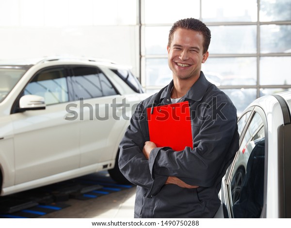 a\
professional mechanical employee of a car service workshop stand\
with a red clipboard in his typical work wear in the garage with a\
white car in the background and smile very\
attractive