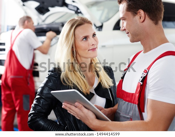 a professional mechanical employee have a sales\
talk about the repairs and a clipboard with the blonde female\
customer and his colleauge make some repairs at the car in the\
background and she smile