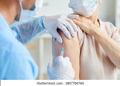 Professional medical worker giving injection to unrecognizable patient sitting next to him into shoulder, over-the-shoulder shot