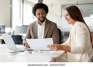 A professional meeting between a man and a woman, both dressed in business attire, discussing documents over a laptop in a bright office setting. - Powered by Shutterstock