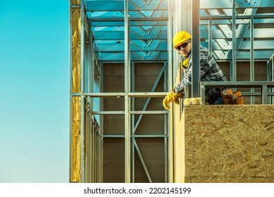 Professional Middle Aged Caucasian Contractor Standing On The Top Of Covered With Wooden Boards Steel House Frame Structure. Clear Blue Sky In The Background.