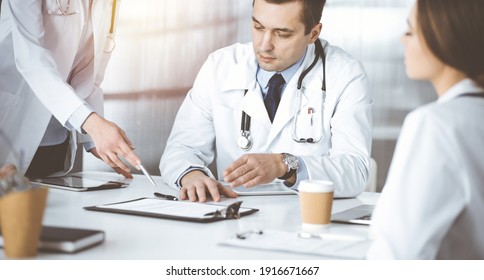 Der professionelle Arzt mittleren Alters spricht mit seinen Kollegen, während er in einem sonnigen Kabinett im Krankenhaus am Schreibtisch sitzt. Ärzteteam, das die Ergebnisse der Blutprobe in einer Klinik diskutiert. Perfekte Medizin