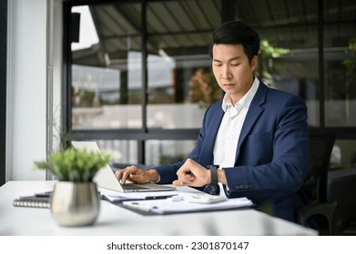 Professional millennial Asian businessman in formal business suit checking time on his wristwatch while working at his desk in the office. - Powered by Shutterstock