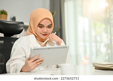 Professional millennial Asian Muslim businesswoman wearing hijab using her digital tablet, thinking and planning a marketing plan, focusing on her business marketing project. - Powered by Shutterstock