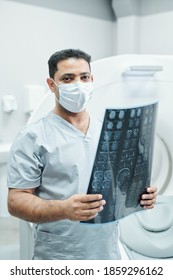 Professional Mixed-race Radiologist In Mask And Uniform Holding X-ray Image Of Patient Head Against Environment Of Medical Office