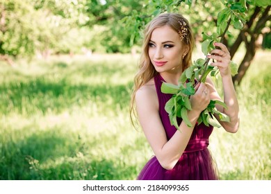 Professional Model With Makeup And Hairstyle Poses In A Green Garden And Holds An Apple Tree Branch In A Fashionable Dress. Beauty Shot And Copy Space.