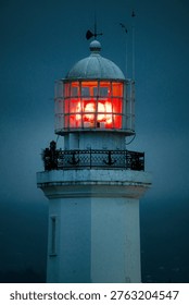 A professional night shot of the upper section of a white lighthouse. The image captures the powerful glowing beacon behind a glass enclosure and metal cage, featuring an ornamental railing with anchor motifs against a moody, dark blue twilight.