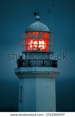 A professional night shot of the upper section of a white lighthouse. The image captures the powerful glowing beacon behind a glass enclosure and metal cage, featuring an ornamental railing with anchor motifs against a moody, dark blue twilight.