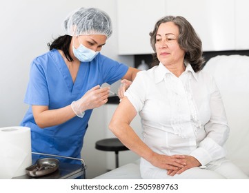 Professional nurse in blue uniform and protective face mask giving antiviral injection to aged woman in medical office. Vaccination, immunization and disease prevention concept - Powered by Shutterstock