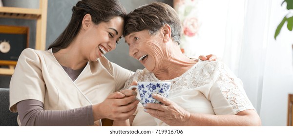 Professional nurse and smiling senior woman laughing while sitting in common room and drinking tea