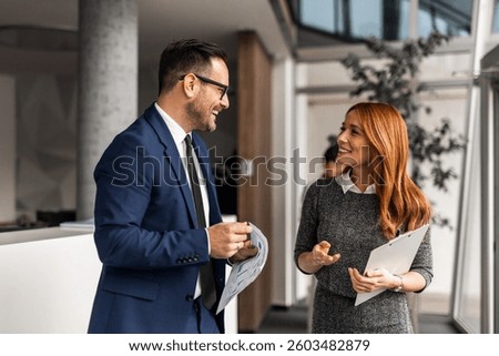 A professional office setting showcasing two colleagues discussing work, sharing ideas, and smiling. They are engaged in a positive interaction, collaboration, and a productive work environment.