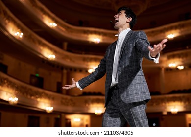 cantante de ópera profesional con elegante atuendo clásico, cantando con la boca abierta en gesto de micrófono con la mano parada en el escenario del teatro. Un hombre caucásico guapo durante el desempeño