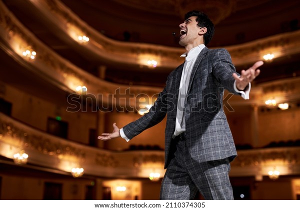 professional\
opera singer in elegant classic outfit, singing hit with open mouth\
in microphone gesture with hand standing on stage in theater.\
Handsome caucasian guy during\
performance