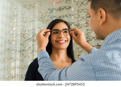 Professional optician fits eyeglasses on a smiling female patient at an optical shop, with various frames displayed in the background - Powered by Shutterstock