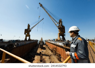 Professional overseeing extensive shipbuilding process, massive cranes erect vessel in dry dock against a clear sky. Maritime engineer in hardhat and reflective vest monitors construction.