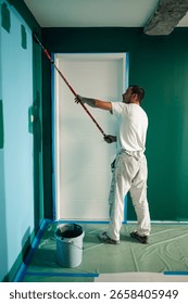 Professional painter using a telescopic paint roller painting a wall green in a house during renovation works
