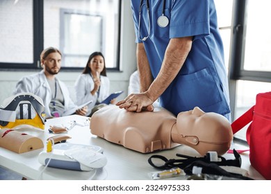 professional paramedic in blue uniform doing chest compressions on CPR manikin near medical equipment and young multiethnic students on blurred background, practicing life-saving skills concept - Powered by Shutterstock