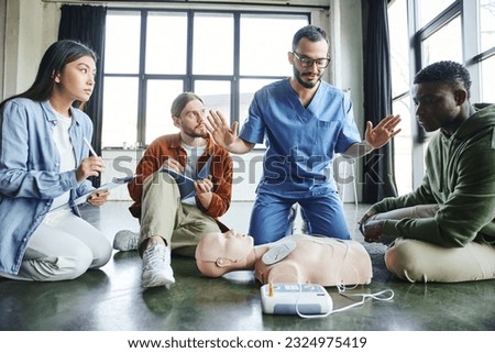 professional paramedic gesturing and talking to multiethnic participants near CPR manikin and defibrillator during first aid seminar in training room, health care and life-saving techniques concept