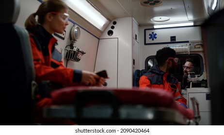 Professional Paramedics Driving On Traffic Accident In Emergency Car. Multi Ethnic Medical Team Talking In Ambulance Van. Smiling Woman In Uniform Browsing Internet On Mobile Phone. First Aid Concept