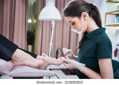 Professional Pedicurist Filing Woman Toenail With Nail File Emery Board. Female Client With Bare Feet Lying On Salon Chair While Nail Specialist Doing Pedicure In Protective Face Mask.